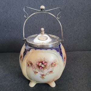 Peut inclure: Un sucrier vintage en céramique crème avec un couvercle et une poignée argentés. Le sucrier présente un motif floral avec des fleurs roses et jaunes. Le bord est orné d'une bande bleu foncé. Le sucrier a trois petits pieds.