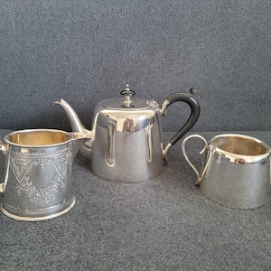 May include: A silver tea set, including a teapot with a black handle, a creamer, and a sugar bowl. The teapot has a decorative finial on the lid. The creamer has an ornate design. The set is displayed on a gray surface.