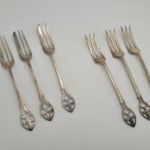 Pode incluir: Seis garfos prateados, cada um com três dentes, dispostos sobre uma superfície branca. Os garfos apresentam desenhos ornamentais vazados na base dos cabos. Provavelmente vintage ou antigos.