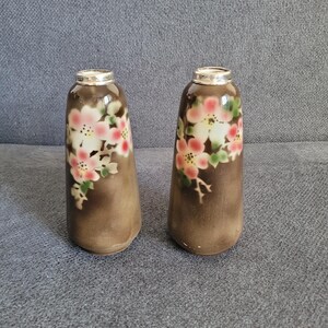 Peut inclure: Deux vases en céramique brune avec un motif floral. Les vases ont un bord argenté et sont décorés de fleurs roses, blanches et vertes.