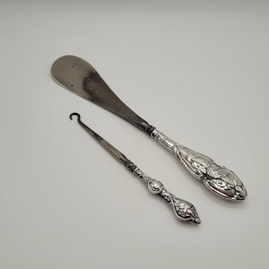 Peut inclure: Deux outils anciens en argent, un chausse-pied et un tire-bouton, sont présentés sur fond blanc. Le chausse-pied a une large lame incurvée et un manche décoratif. Le tire-bouton est fin avec une extrémité en crochet incurvé et une poignée ornée.