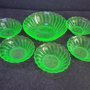 Könnte beinhalten: Ein Set aus sechs grünen Glasschalen. Die Schalen haben ein Spiralmuster an den Seiten und ein kreisförmiges Muster in der Mitte. Sie sind auf einer dunkelblauen Oberfläche angeordnet.