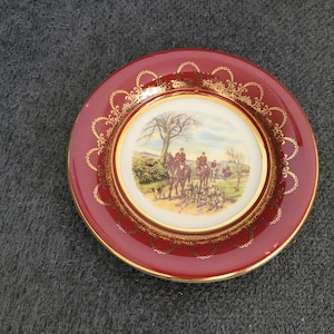May include: A decorative plate with a burgundy rim and a gold-colored filigree border. The center features a pastoral scene with riders on horseback, dogs, and a tree. The plate has a gold-colored rim.