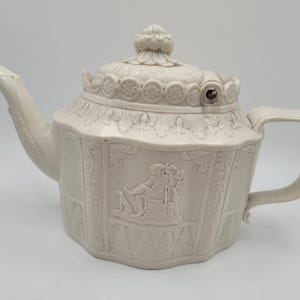 Może przedstawiać: Kremowobiały ceramiczny czajniczek z reliefowym wzorem przedstawiającym siedzącą postać z psem. Czajniczek posiada pokrywkę z kwiatowym uchwytem, zakrzywioną wylewkę oraz ucho. Korpus jest ozdobiony pionowymi panelami z motywami liści.