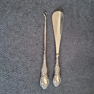Puede incluir: Dos accesorios de zapatos antiguos plateados. Uno es un calzador largo y delgado con un extremo curvo, y el otro es un gancho para zapatos. Ambos tienen mangos ornamentados y detallados con un diseño decorativo.