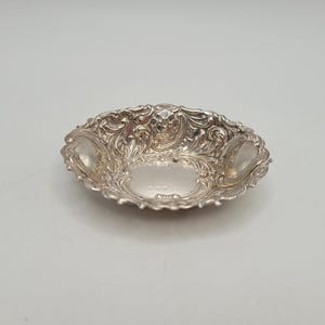 Peut inclure: Un petit plat argenté orné d'un motif tourbillonnant détaillé. Le plat a une forme ovale avec un bord festonné et deux petites sections ovales surélevées de chaque côté. L'intérieur est lisse et réfléchissant.