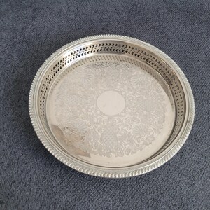 Pode incluir: Uma bandeja redonda prateada com um padrão floral decorativo no centro. A bandeja tem uma borda perfurada e uma borda em forma de corda. A bandeja é feita de metal e provavelmente é usada para servir alimentos ou bebidas.