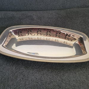 Può includere: Un vassoio rettangolare color argento con un bordo decorativo bordato. Il centro presenta un design traforato di viti. Il vassoio è probabilmente in metallo ed è adatto per servire cibo o esporre oggetti.