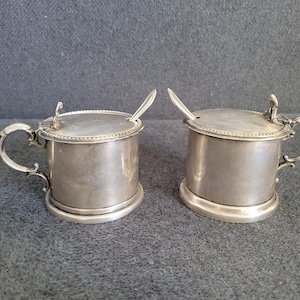 May include: Two silver-colored lidded containers with small spoons. Each container has a handle and a decorative finial on the lid. The containers are cylindrical with a slightly flared base, and are likely for serving condiments.