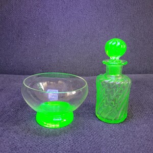 Könnte beinhalten: Eine klare Glasschale und eine kleine grüne Glasflasche mit einem Verschluss. Die Schale hat eine klare Oberseite und einen grünen Boden. Die Flasche hat ein strukturiertes Design und einen runden Verschluss. Die Objekte befinden sich auf einer dunkelblauen Oberfläche.
