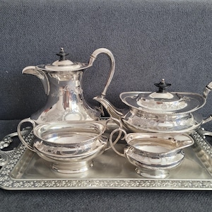 May include: A silver-plated tea service set comprising a tall coffee pot, a teapot, a sugar bowl and a creamer, all featuring ornate engraved patterns and black finials. The four pieces are arranged on a rectangular silver-plated serving tray.