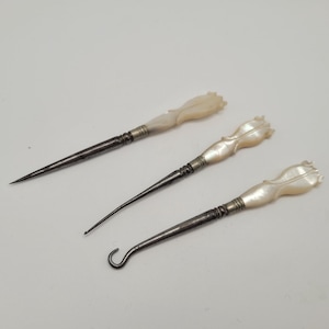 May include: Three antique tools with iridescent mother-of-pearl handles shaped like flower buds. Each tool features a metallic shaft connected to the handle by a silver-toned ferrule. The set includes a pointed pick, a curved hook, and a button hook.