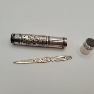 Op de afbeelding: Een zilveren naaigerei met een bloemmotief. De set bevat een cilindrische koker, een kleine vingerhoed en een klein mesachtig gereedschap. De koker is ongeveer 7.6 cm lang.