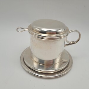 Könnte beinhalten: Ein silberfarbener vietnamesischer Kaffeefilter aus Metall, bestehend aus einer zylindrischen Kammer, einem runden Deckel mit kleinem Schlaufengriff und einer passenden Untertasse. Das Gerät verfügt über einen seitlichen Griff und eine polierte Oberfläche.