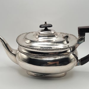 Könnte beinhalten: Eine silberne Teekanne mit einem dunklen Holzgriff und Knauf. Die Teekanne hat einen abgerundeten Körper, einen gebogenen Ausguss und einen dekorativen, perlenbesetzten Rand um den Deckel. Der Griff hat eine dunkle, geschwungene Form.