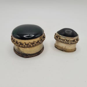 Pode incluir: Duas pequenas caixas antigas com tampas arredondadas verde-escuras. As caixas têm uma base de metal dourado com uma borda decorativa de contas. A caixa menor está ao lado da maior.