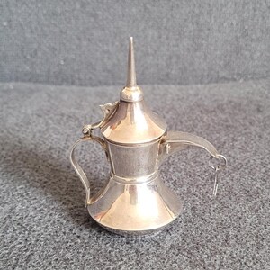 Peut inclure: Une théière miniature en argent avec un couvercle pointu et un bec verseur à charnière. La théière a une petite poignée et une chaîne attachée au bec.