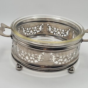 Op de afbeelding: Een ronde zilverkleurige metalen serveerschaal met een ingewikkeld opengewerkt filigraanpatroon, voorzien van twee zijhandgrepen en drie kleine bolvormige voetjes. Een heldere glazen inzetstuk zit in het metalen frame als een kom.