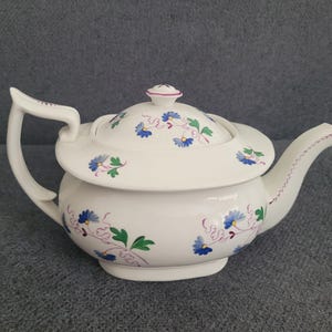 Könnte beinhalten: Weiße Keramik-Teekanne mit Deckel, verziert mit blauen Blumenmustern und grünen Blättern. Die Teekanne hat einen gebogenen Griff und einen Ausguss. Der Deckel ist mit einem kleinen rosa Knauf versehen. Für Tee.