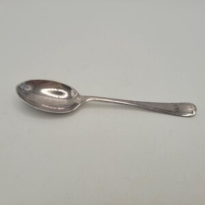 Puede incluir: Una cuchara de plata con un cuenco redondeado y un mango delgado. El mango tiene un extremo decorativo con la palabra "ONE" grabada. La cuchara está sobre un fondo liso y claro.