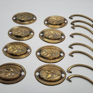 Może przedstawiać: Zestaw antycznych mosiężnych uchwytów i gałek do szuflad. Owalne uchwyty mają podniesiony wzór liści i żołędzi. Uchwyty są zakrzywione o prostym designie. Idealne do renowacji mebli.