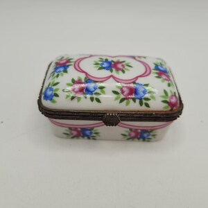 May include: A rectangular porcelain trinket box with a hinged lid. The box is white with a floral pattern of pink and blue flowers, green leaves, and pink ribbon accents. The edges are trimmed with a bronze-coloured metal.