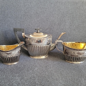 Op de afbeelding: Een zilveren theeservies bestaande uit een theepot, een melkkan en een suikerpot. Elk stuk is voorzien van een geribbelde voet, gedetailleerde gravures en een goudkleurige binnenkant. De theepot heeft een donker handvat en een deksel.