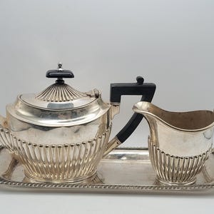 May include: A silver-plated tea set consisting of a teapot with a black handle and finial, a matching creamer, and a rectangular serving tray. Each piece features a fluted lower body design and a decorative rope-style border on the tray edge.