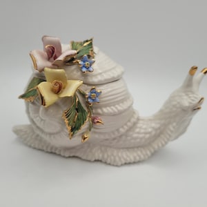 May include: A white ceramic snail figurine with a textured shell and body. The snail is adorned with pink, yellow, and blue floral accents with gold detailing. The figurine is a decorative item.