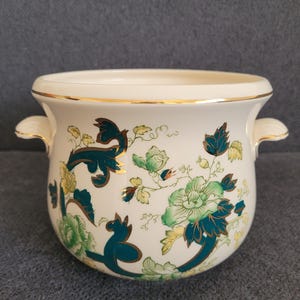 May include: Cream ceramic planter with gold trim and handles. The planter features teal and green floral patterns. A decorative item for home decor.