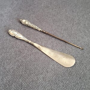 Könnte beinhalten: Zwei silberne Schuhlöffel mit verzierten Griffen. Der größere Löffel hat eine abgerundete Klinge, während der kleinere Löffel eine spitze Spitze hat.