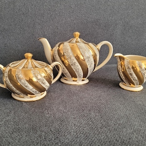 Può includere: Un set da tè di tre pezzi composto da una teiera, una zuccheriera con coperchio e una lattiera. Ogni pezzo presenta un motivo a spirale con bande dorate metallizzate alternate a sezioni bianche decorate con un delicato disegno floreale grigio.