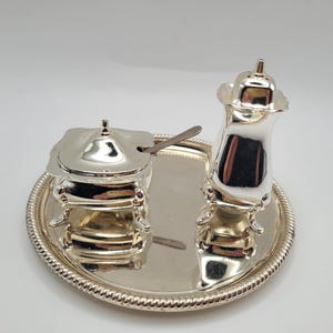 Peut inclure: Un ensemble à condiments argenté comprenant un petit sucrier carré avec couvercle et cuillère assortie, ainsi qu'une grande salière ou poivrière couverte. Les deux pièces reposent sur des pieds ornés sur un plateau rond à bord perlé.