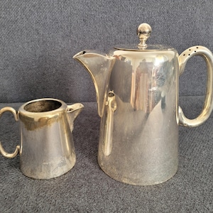 Op de afbeelding: Een zilverkleurige theeset, bestaande uit een theepot met deksel en een melkkan. De theepot heeft een gebogen handvat en een tuit. De melkkan heeft een klein handvat en een tuit. Beide stukken zijn van metaal.