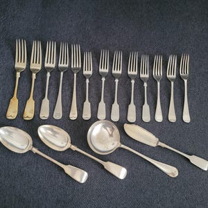 May include: An assortment of antique silverware, including forks, spoons, a ladle, and a butter knife, displayed on a dark blue surface. The forks have tines, and the spoons have oval bowls. The silverware is made of silver and gold.