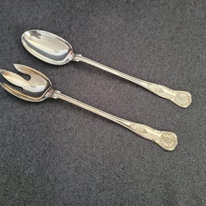 Puede incluir: Dos utensilios de servir plateados: una cuchara y un tenedor. La cuchara tiene un cuenco ovalado y el tenedor tiene tres púas. Ambos tienen mangos largos con un diseño floral decorativo en el extremo. Sobre una superficie gris oscuro.