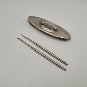 Könnte beinhalten: Ein ovales silbernes Etui mit einem texturierten guillochierten Muster und einem zentralen Metallverschluss, begleitet von zwei passenden silbernen Werkzeugen mit schlanken, spitzen Enden und texturierten Griffen, alle mit sichtbaren Punzen.