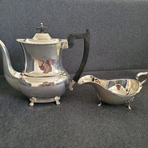 Op de afbeelding: Een zilveren theeservies met een theepot en een melkkan. De theepot heeft een zwarte handgreep en een deksel met een kleine decoratieve knop. De melkkan heeft een gebogen tuit en een klein handvat. Beide stukken staan op kleine voetjes.