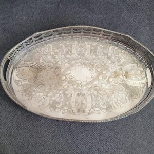 Può includere: Un vassoio da portata ovale color argento caratterizzato da un intricato motivo floreale e a volute inciso sulla superficie centrale. Il vassoio presenta un bordo rialzato traforato con un design a griglia e due manici integrati.