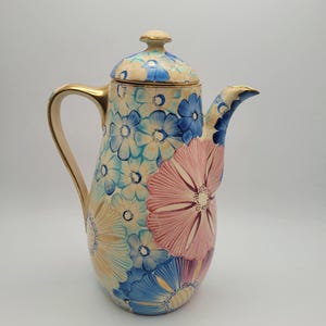 Pode incluir: Uma cafeteira alta de cerâmica com tampa, alça e bico. A cafeteira é decorada com grandes desenhos florais coloridos em tons de azul, rosa e amarelo. A alça e o bico têm detalhes dourados.