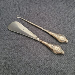 Peut inclure: Deux chausse-pieds et un crochet à boutons en argent antique sur une surface grise. Le chausse-pieds a une lame large et incurvée. Le crochet à boutons a une extrémité crochetée. Les deux ont des manches texturés avec un motif ovale.