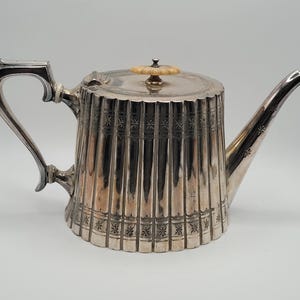 Op de afbeelding: Zilveren theepot met een gegroefde body en een gebogen handvat. De theepot heeft een lange tuit en een deksel met een decoratieve knop. De body is versierd met geëtste sterpatronen. Een klassiek stuk voor de theeservice.