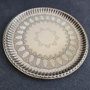 May include: A round silver-toned metal serving tray featuring a polished center, a decorative engraved border with repeating leaf-like patterns, and a scalloped, fluted edge. The tray has a reflective surface and a consistent metallic finish.