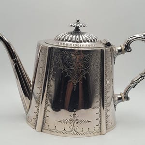 Op de afbeelding: Een zilverkleurige metalen theepot met een achthoekig lichaam voorzien van gedetailleerde bloem- en krulmotieven. De pot heeft een gebogen tuit, een decoratief handvat met witte isolatoren en een afneembaar deksel met een kleine knop.