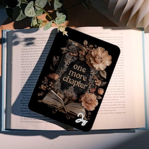 May include: A black bookmark featuring the text "one more chapter" in gold lettering, framed by floral designs. The bookmark is placed on an open book with pale blue pages, with a plant in the background.