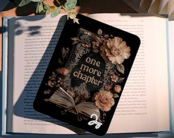 May include: A black bookmark with the words "one more chapter" in gold script, surrounded by floral illustrations. The bookmark is resting on an open book with light blue pages, with a sprig of greenery in the background.