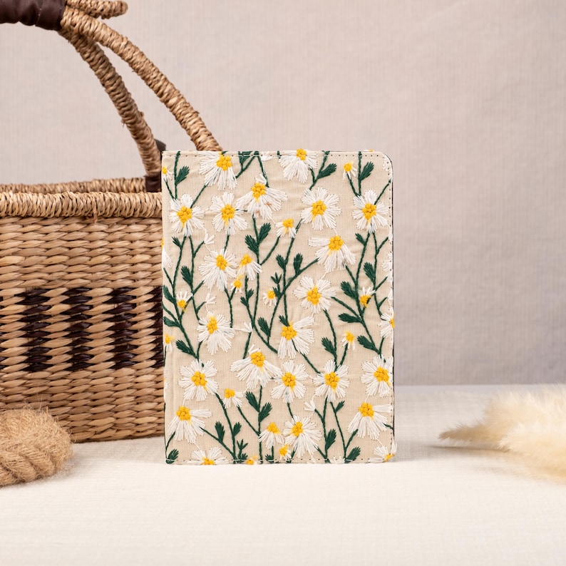 May include: A beige linen notebook cover with a white daisy floral embroidery pattern. The cover has a subtle texture and a stitched border.