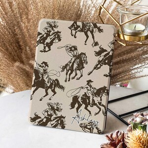 Puede incluir: Una funda para tablet beige con un patrón repetido de siluetas marrones de mujeres montando a caballo. El texto "Ashley" está impreso en cursiva en la funda.