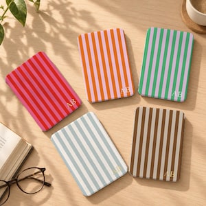 Pode incluir: Cinco cadernos retangulares com riscas verticais em vermelho e rosa, laranja e rosa, verde e rosa, azul claro e branco, e castanho e branco. Cada caderno apresenta o monograma dourado AB impresso no canto inferior direito.
