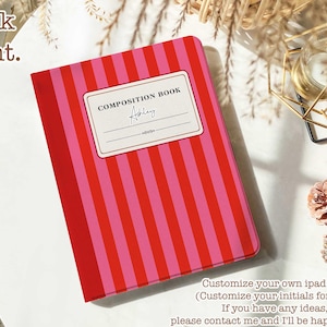 Puede incluir: Un cuaderno de composición a rayas rojas y rosas con una etiqueta blanca que dice "COMPOSITION BOOK" y tiene un espacio para escribir un nombre. El nombre "Ashley" está escrito en cursiva en la etiqueta. El cuaderno está sobre una superficie blanca con una superficie tejida marrón y blanca en el fondo.